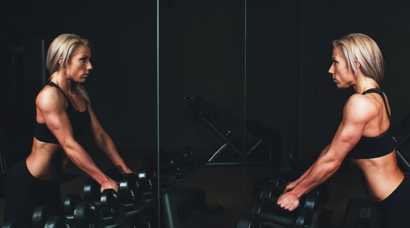 woman wearing black top top holding black dumbbells standing in front of mirror