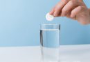 A hand dropping an effervescent tablet into a glass of water on a light blue background.
